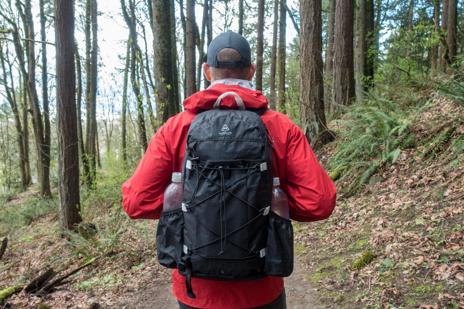 A hiker in a red jacket wearing the Hyperlite Mountain Gear Daybreak on a forest trail with two Smartwater bottles in the side pockets