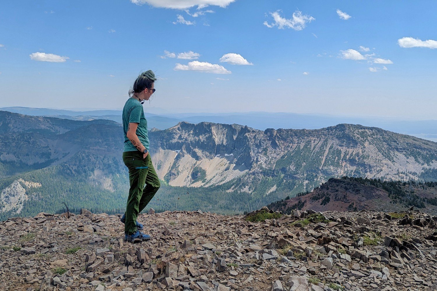A hiker wearing green REI Trailmade Pants on top of a mountain - there's a distant view of more peaks and a blue sky dotted with clouds